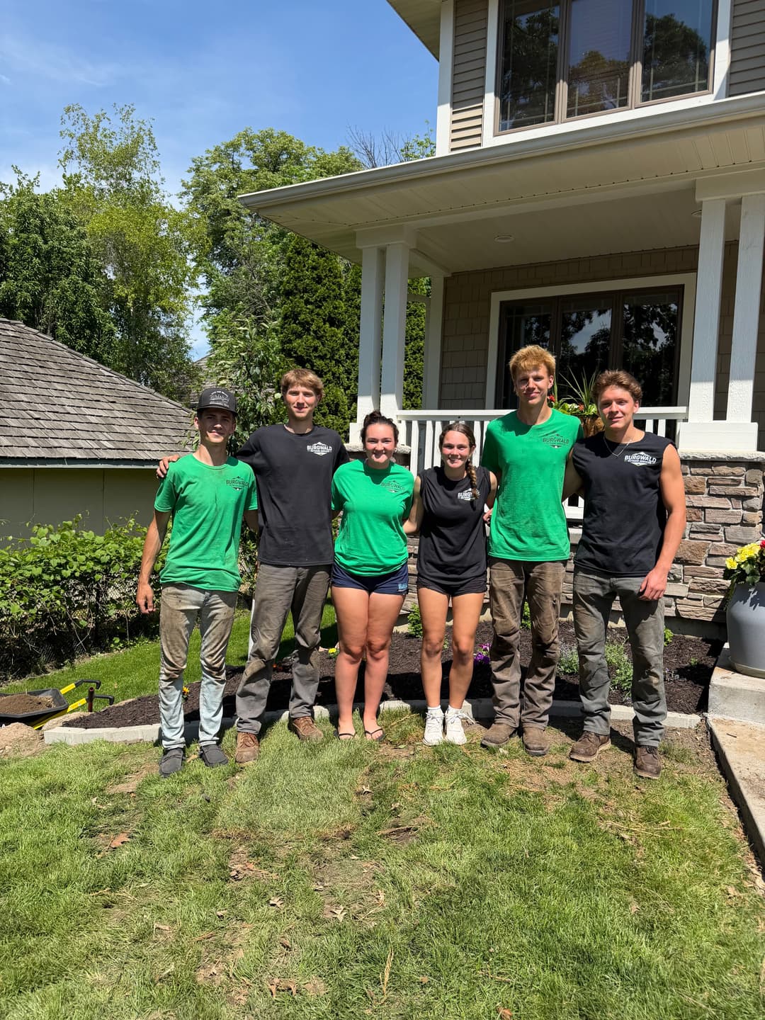 Team of six landscaping workers in green and black shirts pose in front of a house on a sunny day.